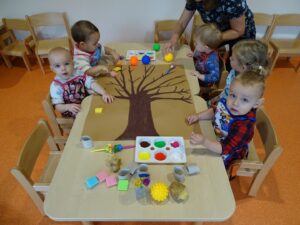 Piątka dzieci siedzi przy stoliku i z pomocą opiekunki rozpoczynają malować drzewko