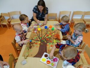 Opiekunka wyjaśnia dzieciom przy stoliku jaki sposób malować farbami drzewko