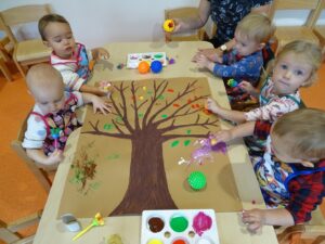 Dzieci siedzą przy stoliku, na któym leży namalowane drzewo, trójka z nich maluje gąbeczkami
