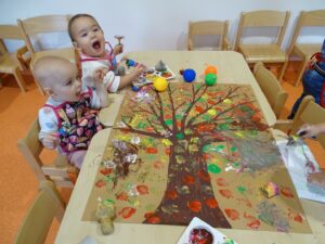 Dwójka dzieci siedzi przy stoliku, na którym leży ukończona praca malowanego jesiennego drzewa