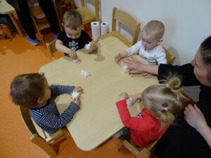 Dzieci siedzą przy stoliku i wkładają wate do kubeczków z pomocą opiekunki