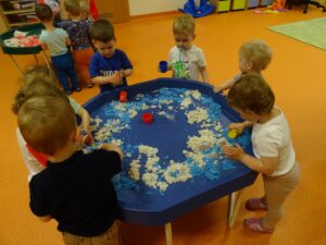 Dzieci stoją przy stoliku i bawią się niebieskim piaskiem